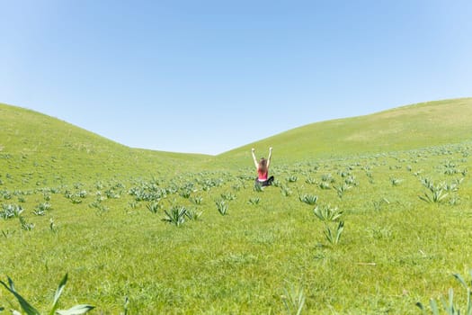 Back view of cheerful women sitting in the field with hands up, green field an blue sky view behind. Faceless.