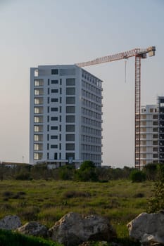 A modern building rises beside a construction crane, surrounded by lush greenery under a soft evening light.