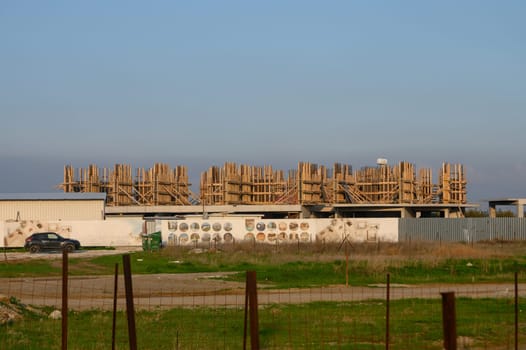 Workers are busy assembling wooden scaffolding on a large construction site surrounded by open fields and buildings.