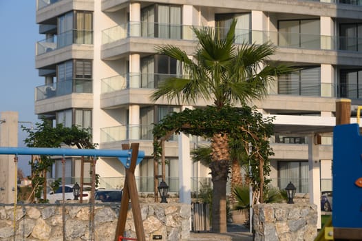 Palm trees sway gently by a modern building, overlooking a tranquil playground while the sun sets on the horizon.