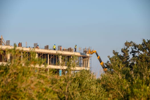 Workers collaborate on a construction site, raising a new structure amidst greenery under the warm sun.