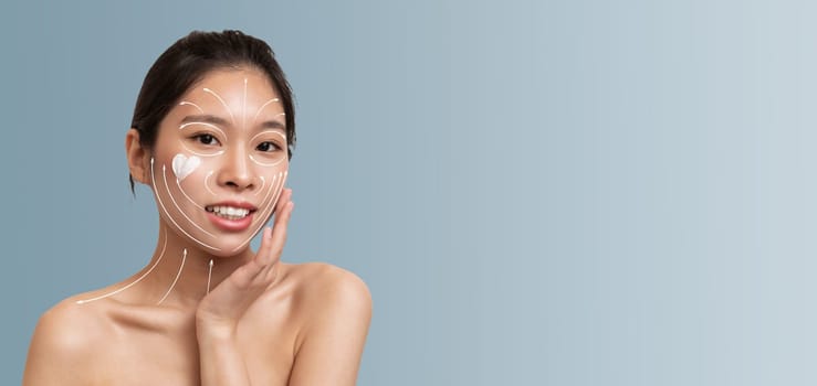 Asian woman enjoys her skincare routine while applying facial cream. She's smiling and appears relaxed, showcasing her commitment to beauty and self-care in a minimalist setting.