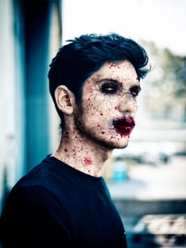 Scary male zombie standing on outdoor in urban setting, with white eyes and bloody mouth, looking at camera