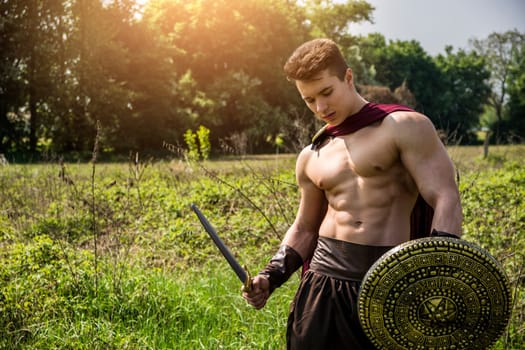 A shirtless muscular young man in a field holding a sword and shield in a roman or spartan, greek ancient soldier costume. Photo of a warrior posing with a sword and shield in a picturesque field