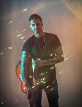 A young handsome man holding a guitar in his right hand in studio shot, surrounded by light sparks. Photo of a man playing a guitar with passion and skill