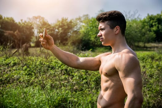 A shirtless muscular young man holding a smartphone in a field to take a selfie. Photo of a shirtless man holding a smartphone in a field