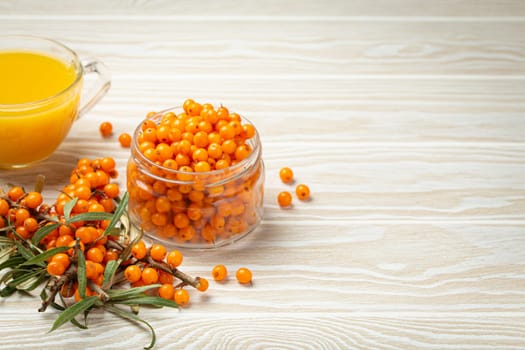 Sea buckthorn healthy hot tea in transparent glass cup, ripe berries in glass jar and branches with leaves on white wooden rustic background, for skin, heart, vessels and immune system. Copy space.