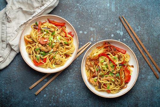 Two bowls with Chow Mein or Lo Mein, traditional Chinese stir fry noodles with meat and vegetables, served with chopsticks top view on rustic blue concrete background.