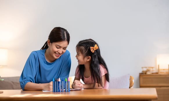 A young family spends their free time together in the living room at home. Mother and little daughter draw pictures with crayons on paper, smiling happy..