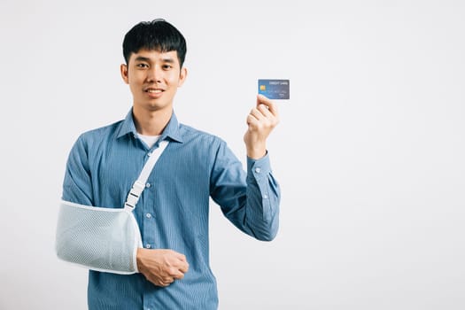 Despite a damaged arm, a man wears a support splint, paying medical bills with a credit card after an accident. Happy Asian man in a sling on white background, emphasizing health care. Copy space