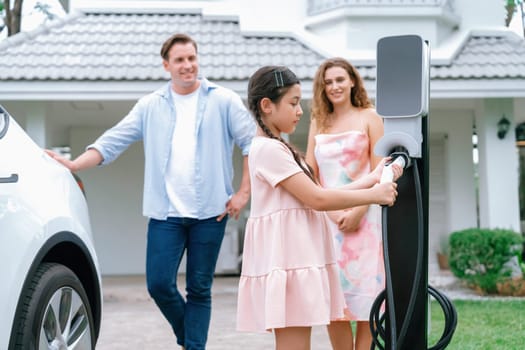 Happy little young girl learn about eco-friendly and energy sustainability as she help her family recharge electric vehicle from home EV charging station. EV car and modern family concept. Synchronos