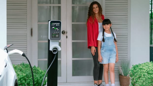 Happy little young girl learn about eco-friendly and energy sustainability as she help her mother recharge electric vehicle from home EV charging station. EV car and modern family. Panorama Synchronos