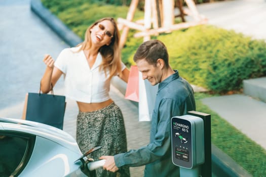 Young couple travel with EV electric car charging in green sustainable city outdoor garden in summer shows urban sustainability lifestyle by green clean rechargeable energy of electric vehicle innards