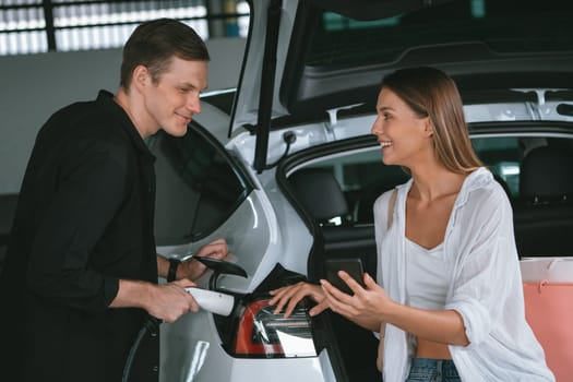 Young couple travel with EV electric car to shopping center parking lot charging in downtown city showing urban sustainability lifestyle by green clean rechargeable energy of electric vehicle innards