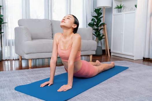 Asian woman in sportswear doing yoga exercise on fitness mat as her home workout training routine. Healthy body care and calm meditation in yoga lifestyle with comfortable and relaxation. Vigorous