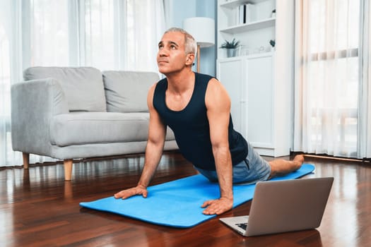 Senior man in sportswear being doing yoga in meditation posture on exercising mat at home. Healthy senior pensioner lifestyle with peaceful mind and serenity. Clout