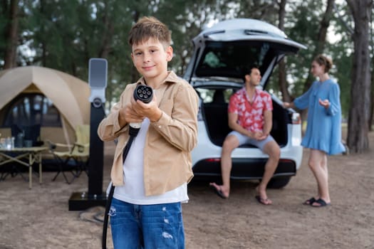 Little boy holding EV charger and point at camera with his family sitting on the trunk in background. Road trip travel with alternative energy charging station for eco-friendly car concept. Perpetual