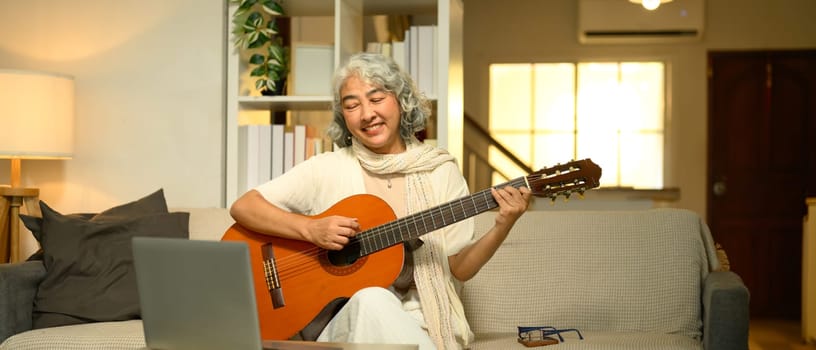 Cheerful elderly woman plays an acoustic guitar while learning to play acoustic guitar online.