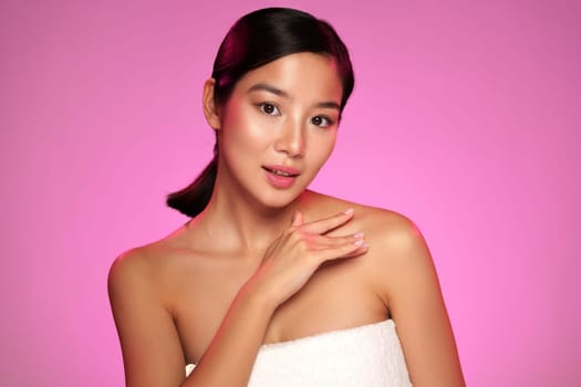 A woman applies skincare products on her shoulder, showcasing her glowing skin. She poses with confidence against a vibrant pink backdrop, highlighting beauty and self-care.