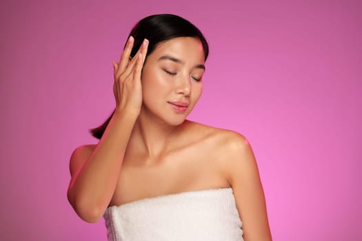 A woman gently touches her hair and closes her eyes, exuding confidence and tranquility. The bright pink background enhances her radiant skin, highlighting beauty and self-care.