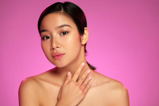 A model poses gracefully with a serene expression, using her hand to accentuate her neck and shoulder in a well-lit studio with a vibrant pink backdrop.