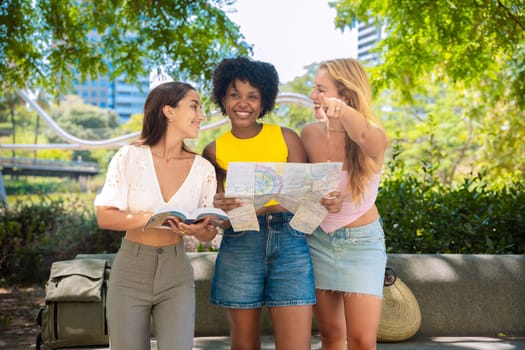 Three happy young women stand outdoors, looking at maps and a travel guide. They smile while planning their trip, enjoying a fun adventure in a sunny urban setting with trees and buildings.