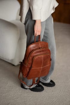 beautiful girl in gray pants and beige sweater posing with a small brown leather backpack.