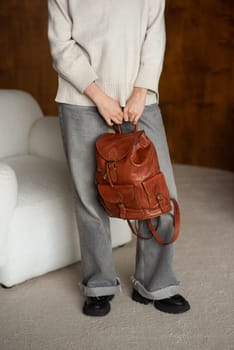 beautiful girl in gray pants and beige sweater posing with a small brown leather backpack.
