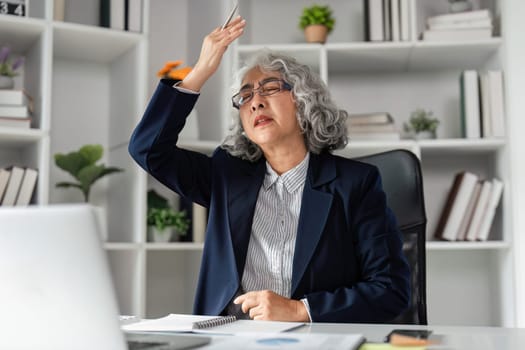 An elderly businesswoman with curly gray hair uses a pen to fan herself, showing signs of exhaustion amidst paperwork and her laptop in a vibrant office environment.