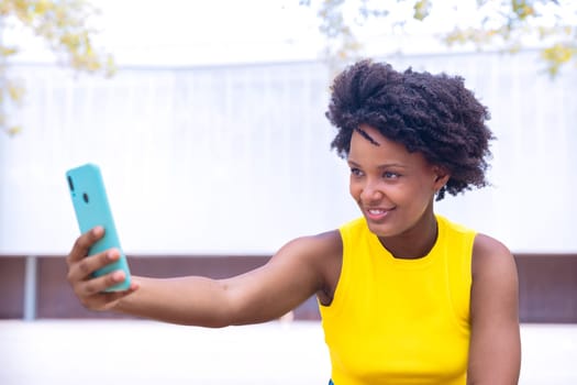 A joyful young woman with curly hair wearing a yellow top takes a selfie outdoors, smiling and gesturing peace. Vibrant scene showcasing happiness, individuality, and modern lifestyle.copy space