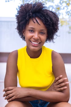 A cheerful young woman with natural curly hair wearing a yellow top smiles confidently while posing outdoors. Bright and vibrant setting, showcasing joy, beauty, and individuality.