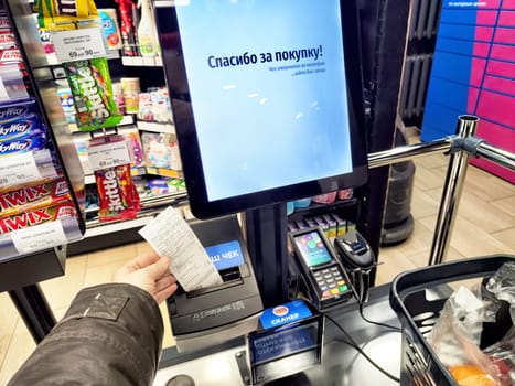 Kirov, Russia - December 11, 2024: A customer completes a transaction at a self-service checkout station, holding the receipt in a modern grocery store setting.