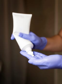 A cosmetologist squeezes cream out of a large tube. Close-up of hands in medical gloves and cream on the palm. The gloves are blue.
