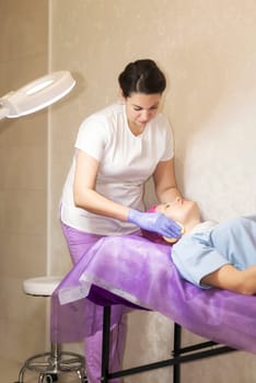 The cosmetologist prepares the client for the procedures. Wipes the face with sponges. The client woman lies on the couch, A bright round lamp is on in the office.