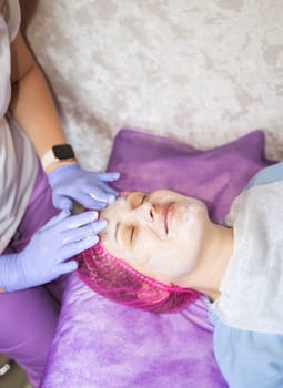 A cosmetologist is performing a facial procedure on a client. The doctor is wearing purple rubber gloves. The cosmetologist is applying cream to a female client's face.