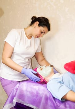A female cosmetologist doctor makes a facial mask for a client. A female client lies on a couch. The cosmetologist removes an alginate mask from her face. facial rejuvenation mask in facial clinic