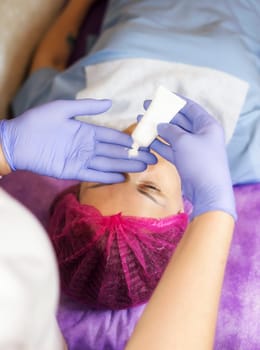 The cosmetologist holds a small tube of cream in her hands. A female client is lying on the couch. The cosmetologist is wearing blue medical gloves. close-up of hands and a tube of cream