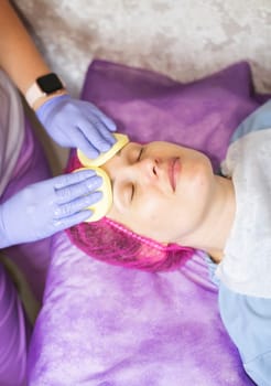A cosmetologist wipes a woman client's face with sponges. The cosmetologist holds two sponges. The client lies on a couch, her hair is tucked into a cap. The colors in the photo are blue and purple