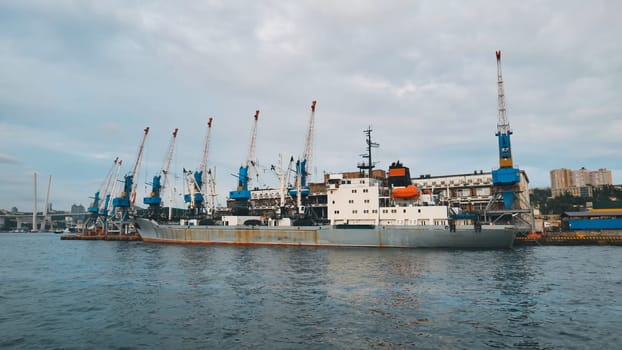 Cargo ship is docking at the commercial sea port of vladivostok, with cranes operating and the zolotoy rog bridge in the background