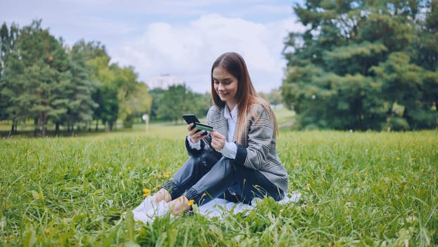 Professional working remotely, processing digital payment via credit card and mobile device while resting on urban park lawn