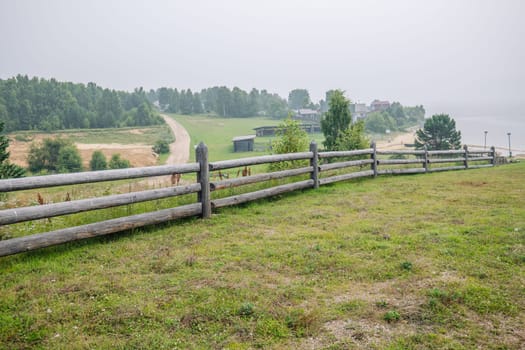 Rural landscape featuring wooden fence, dirt path, misty forest, distant village, winding river during foggy morning light