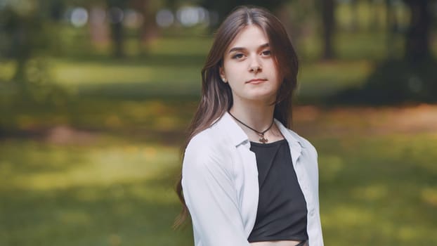 Young woman with long brown hair, dressed in a white shirt and black top, posing confidently in a vibrant city park surrounded by nature