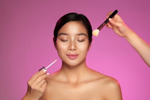 A woman with closed eyes enjoys makeup application as someone uses a brush to apply blush while holding a foundation bottle. The vibrant pink background complements the beauty activity.