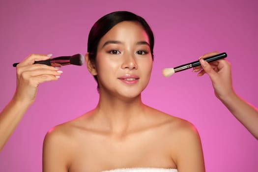 A model sits with her eyes closed as two artists apply makeup to her face, using brushes. The vibrant pink background adds a lively touch to the beauty session in the studio.