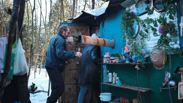 Homeless individuals sheltering in rustic dwelling constructed from salvaged materials, nestled among snow covered winter trees, representing marginalization