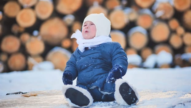 Baby bundled in warm winter clothes, sitting on frozen wood logs and joyfully enjoying a beautiful winter day in the snowy outdoors