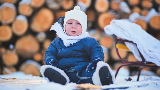 Distressed child bundled in winter gear weeping beside wooden sled, snowy landscape and log pile surrounding small upset toddler
