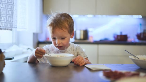 Blond toddler enjoying soup from a white bowl, seated at the kitchen table, immersed in a cozy mealtime experience