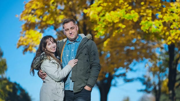 Loving couple walking through golden leaved park, holding each other close during warm autumn afternoon