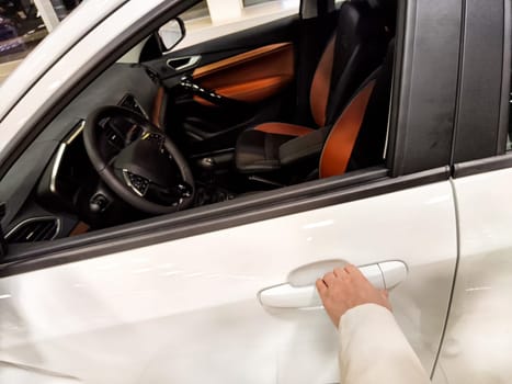 A hand grasping the door handle of a modern white vehicle parked under soft indoor lighting.